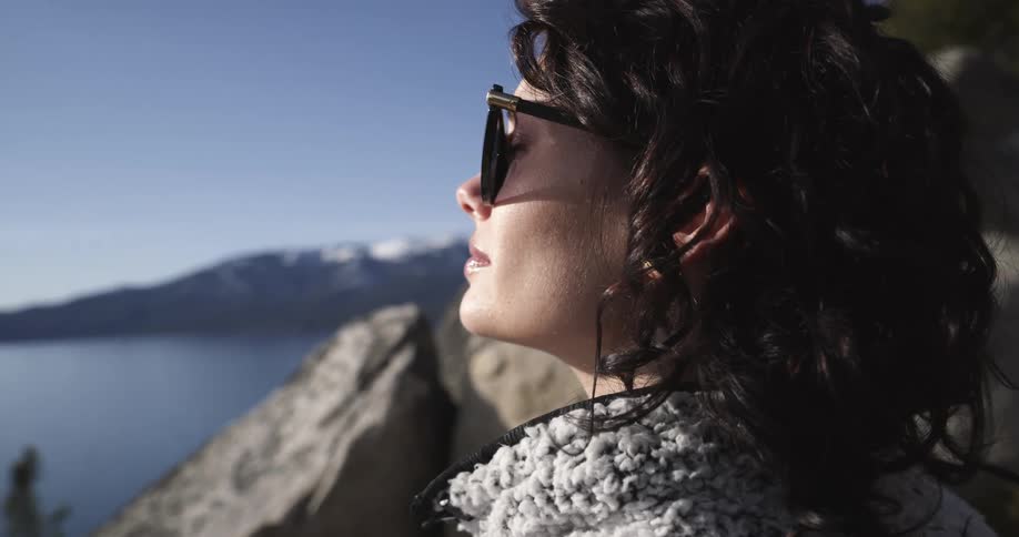 A woman in sunglasses looks out over a lake from a mountain on its shore on a clear day