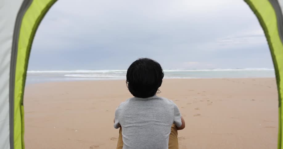 Teenage boy with grey t shirt and brown pants sits in front of a tent on the beach