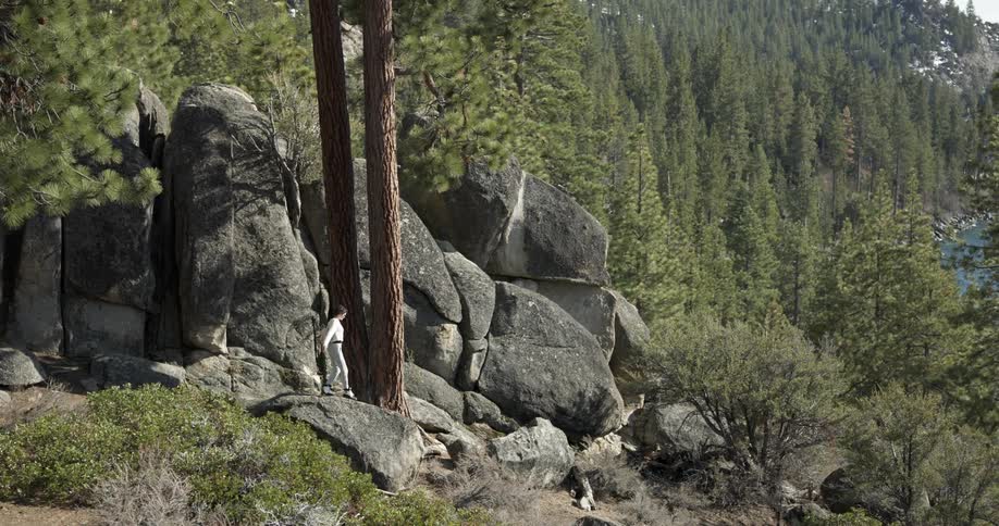Woman climbs onto boulder near forest covered mountain overlooking water