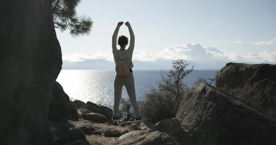 Woman climbs over uneven terrain and claps after reaching shore overlooking water