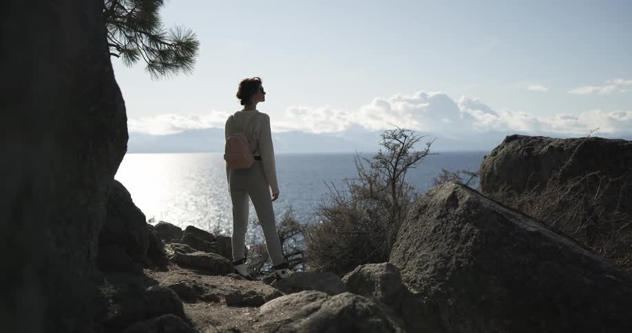 Young woman hiking in the mountains stands atop a rock in near a lake looking at the horizon