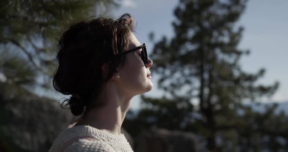 A woman with sunglasses standing near a lake with trees the sky and clouds in the background