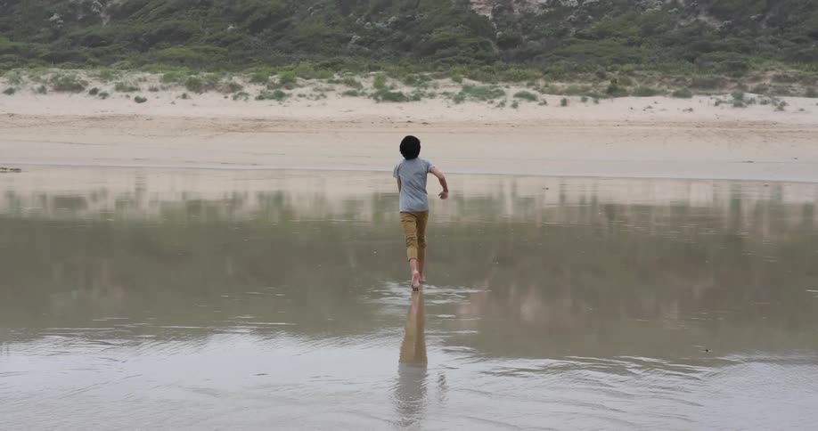 Teenage boy with a grey t shirt and brown pants runs barefoot on the wet sand beach