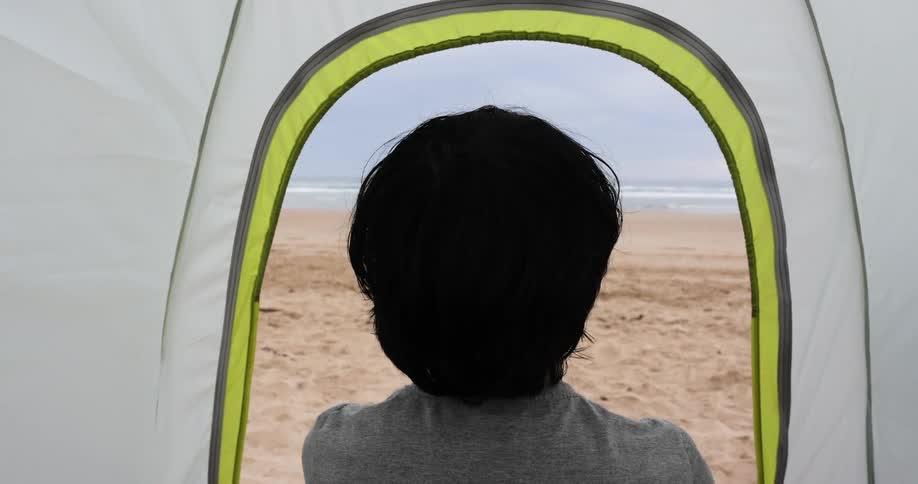 Teenage boy with a grey t shirt sits in a tent on the beach and watches the sea