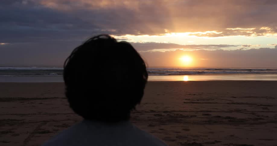 A silhouetted boy watching a sunset on a sandy beach