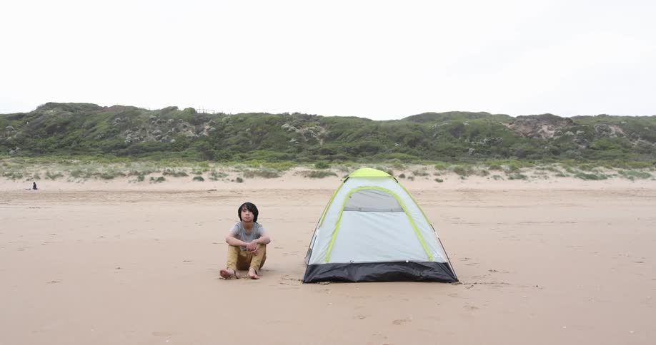 A barefoot boy sitting next to a tent on a sandy beach in the daytime