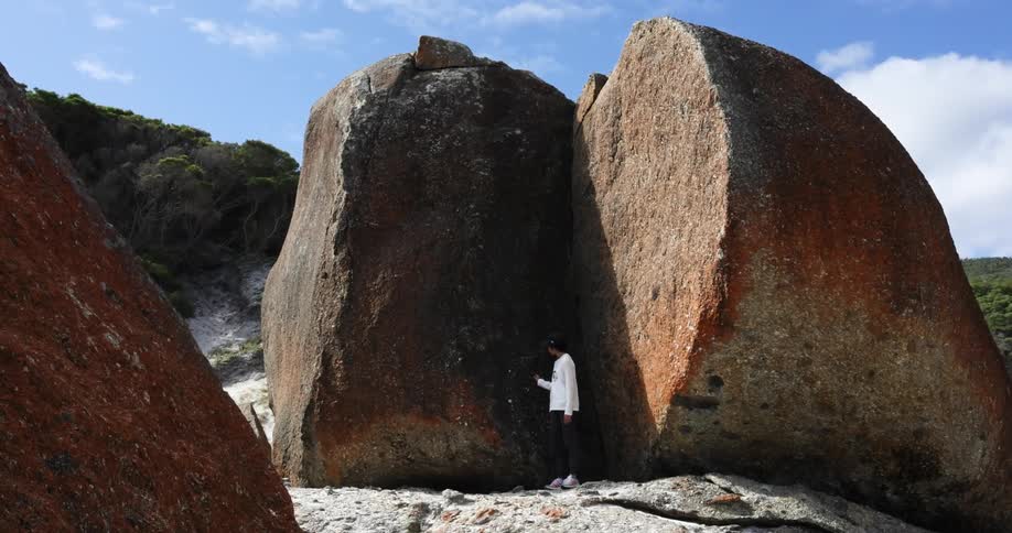 A boy with a hat standing by large boulders with the sky and clouds in the background