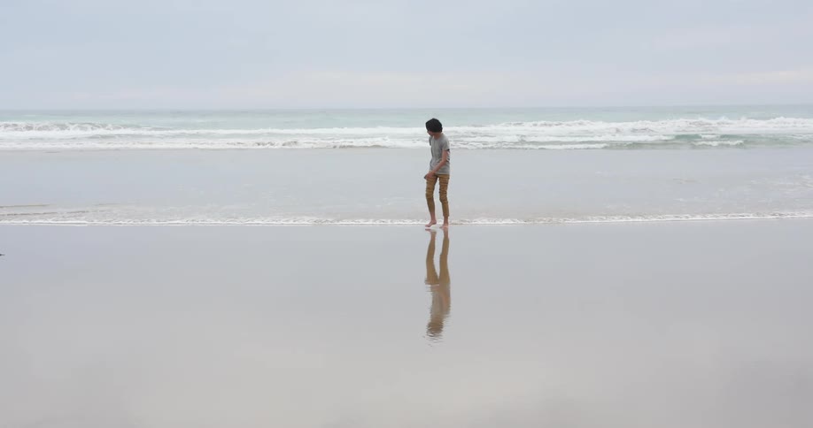 Teenage boy with a grey t shirt and brown pants walks barefoot on the wet sand beach next to the sea