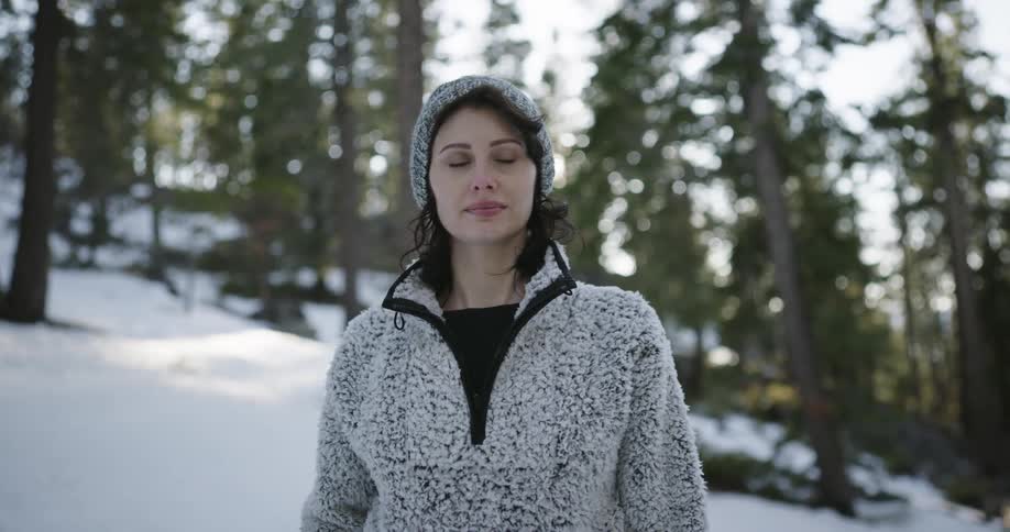 Woman wearing winter clothes stands over the snow in the forest looking at the camera