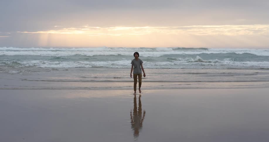 Boy with long hair grey t shirt and brown pants walks on the wet sand beach next to the sea at dusk