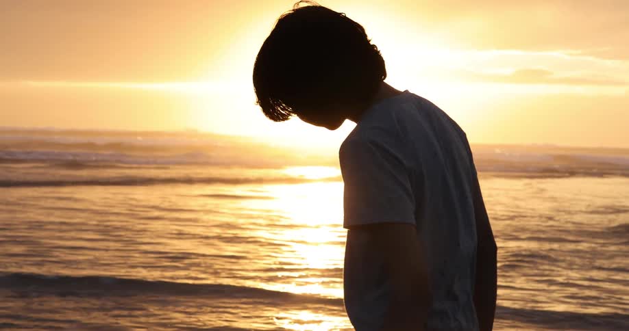 A silhouetted boy standing alone on a beach at sunset