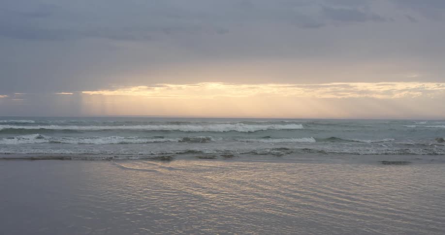 Sunlight peaks through dense clouds as waves crash onto the shore of a beach