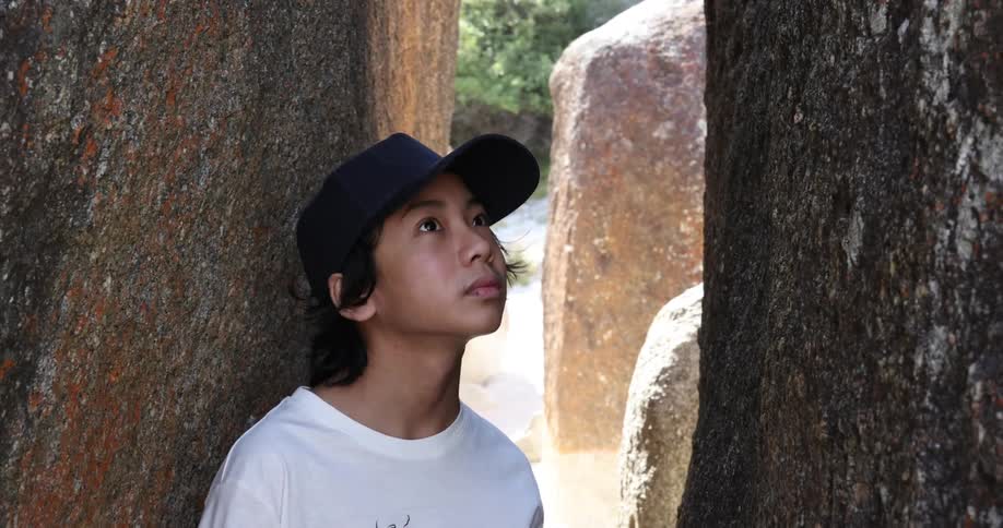 A boy with a hat and a t shirt walking between two rocks with trees in the background