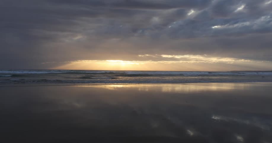 Waves roll in on beach under a cloudy sky with a sunset