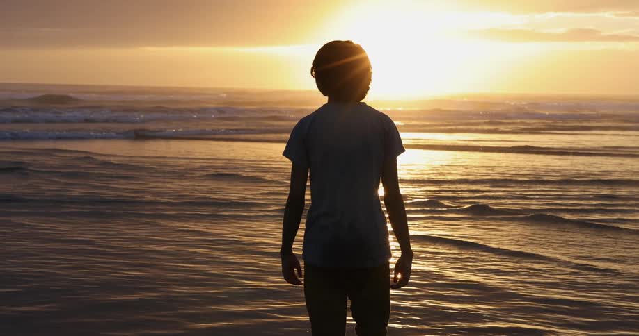 A boy is walking towards the ocean at sunset