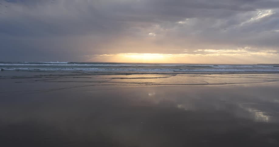 Waves roll in on beach under a cloudy sky with a sunset
