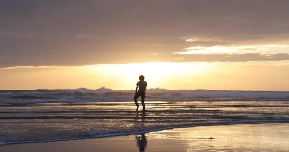 Teenage boy walking barefoot on the wet sand beach is silhouetted against the sunset
