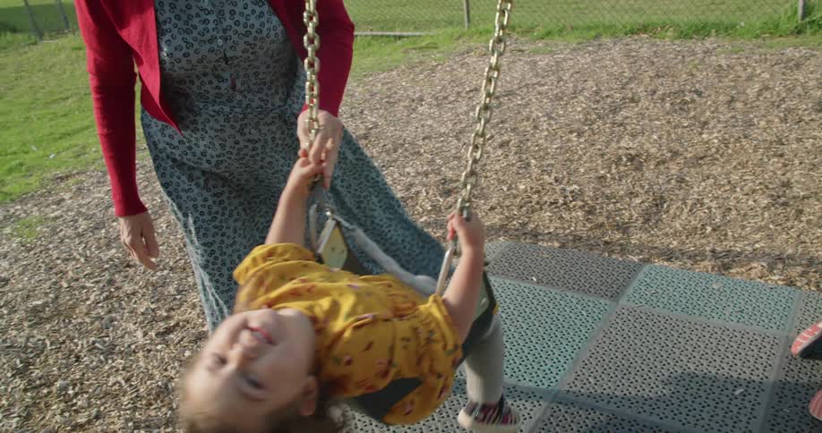 A woman pushing a toddler girl on a playground swing set on a summer day