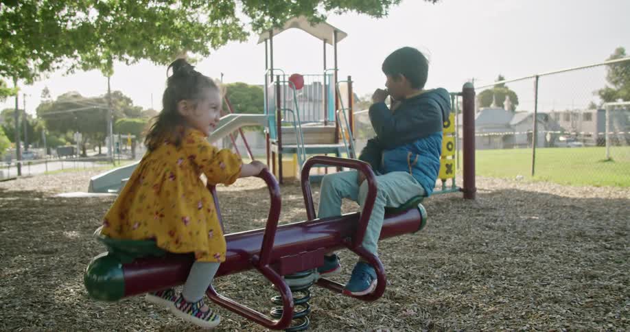 A little girl is smiling and happy playing on the playground seesaw while her brother looks bored