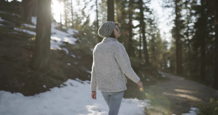 Woman with a woolly hat a grey sweater and jeans walks in the snowy forest