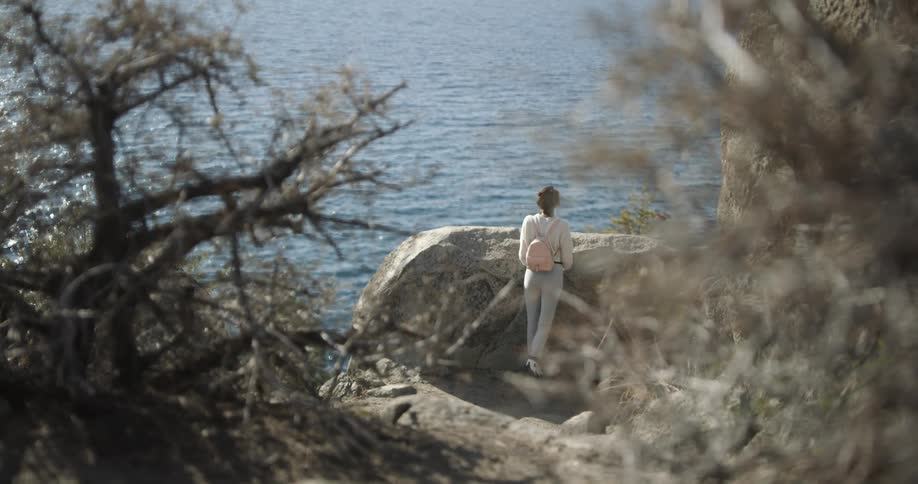 A woman with a backpack and sunglasses by a lake with trees in the foreground