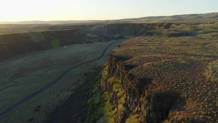 Flying along the edge of a canyon cliff as an empty gravel road winds into the distance