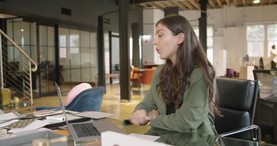 A young woman uses a laptop to participate in a video chat while sitting at a table in an office during the day