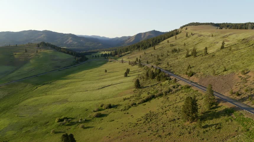 A car travels on a hillside road dotted with trees and a mountain range on the horizon