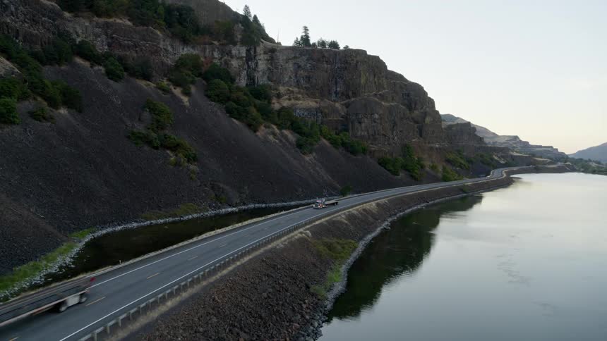 Semi trucks drive on a road between rock cliffs and a lake in the mountains at dawn