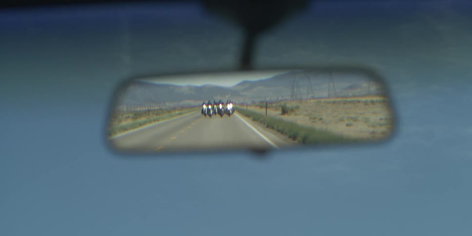 A vehicle with people on motorcycles on the review mirror with the sky and clouds in the background