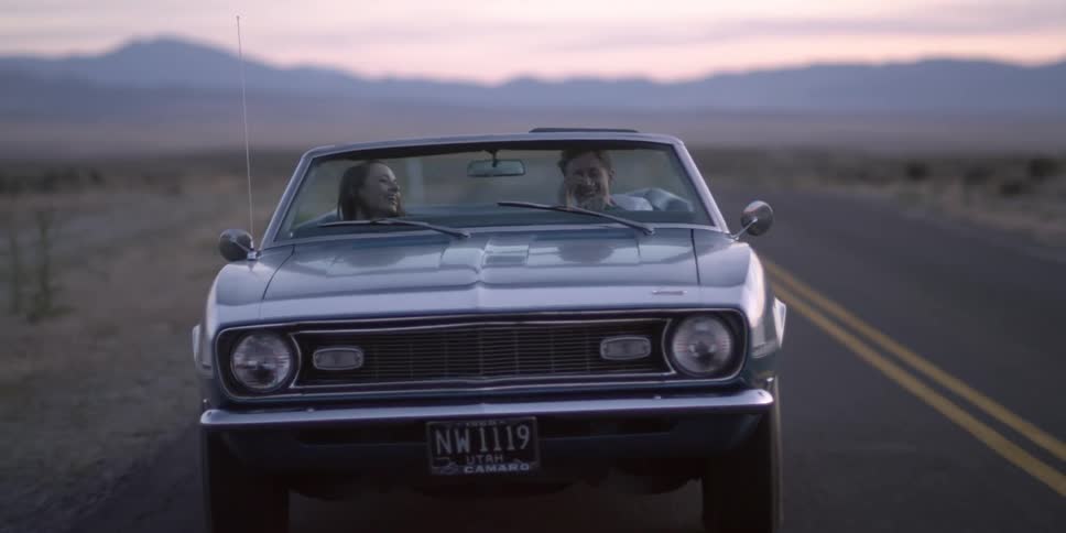 A man and a woman riding in a convertible with the sky and clouds in the background