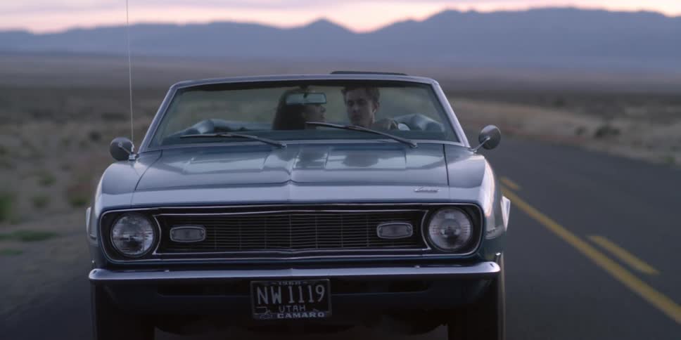 A woman sitting in the passenger seat of a car kisses the driver on the cheek as they travel along a road through a desert in the evening