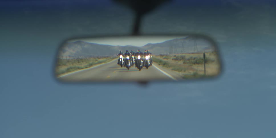A vehicle with people riding motorcycles in a desert in the rearview mirror with the sky and clouds in the background