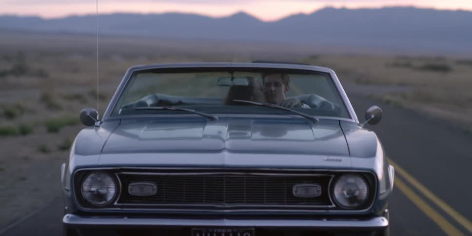 A woman leans across and kisses a man as he drives a convertible on an empty desert highway