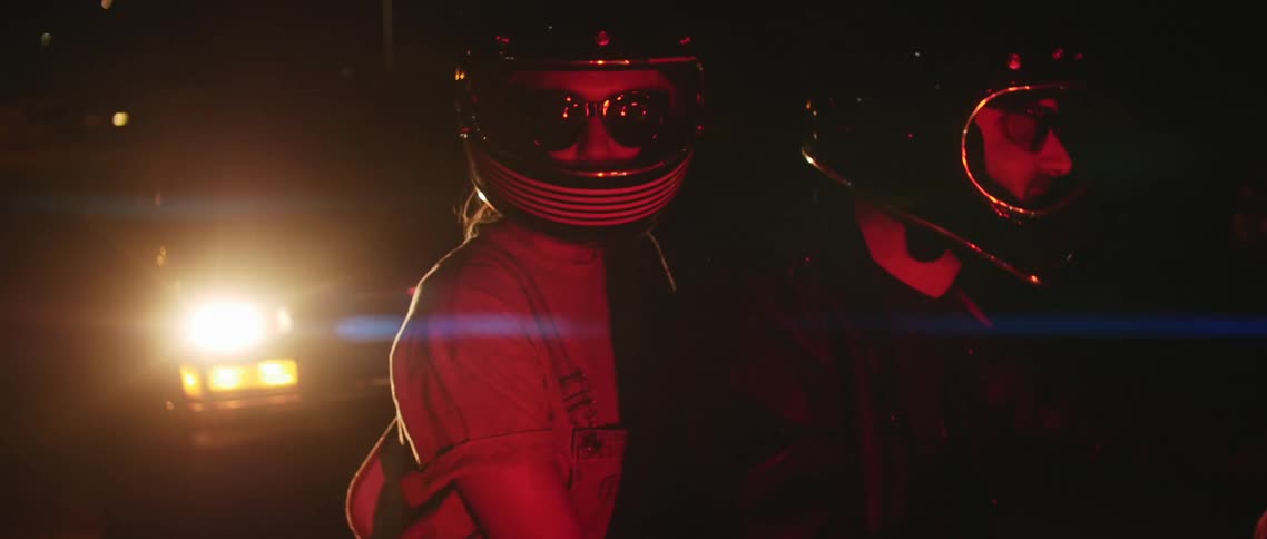 Two people with helmets on a motorcycle with a vehicle with headlights in the background