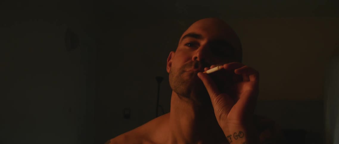Shirtless bald tattooed man smoking a cigarette in a dimly lit room