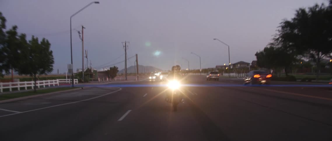 Two people wearing helmets riding a motorcycle in a small towns road with the headlight on at evening