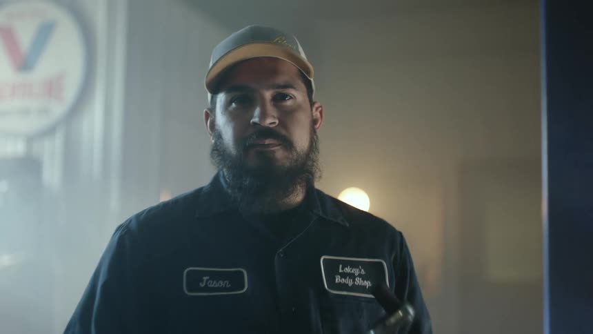 A bearded mechanic holding a wrench in an auto body shop