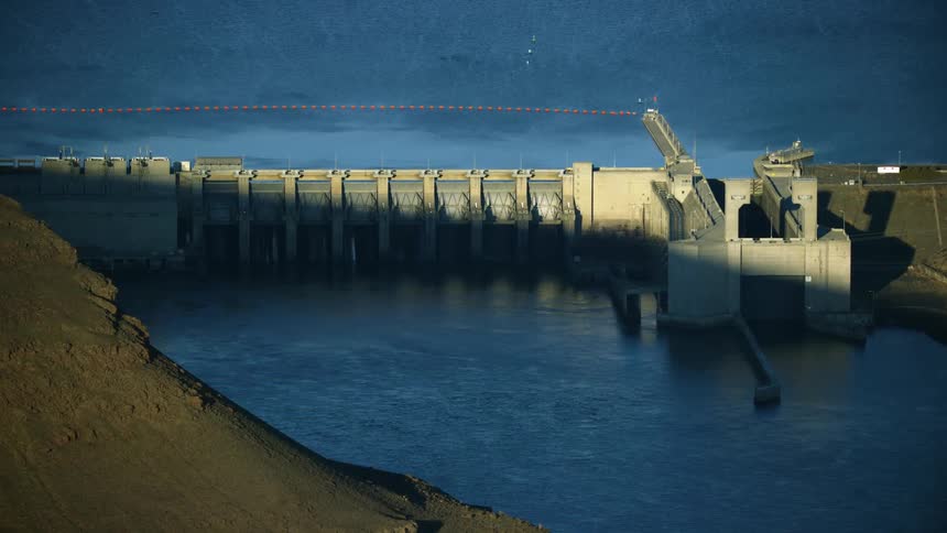 Lower Monumental Dam is revealed from behind a grassy hill in this tight shot aerial communicating that impoundment of the concrete run of river dam