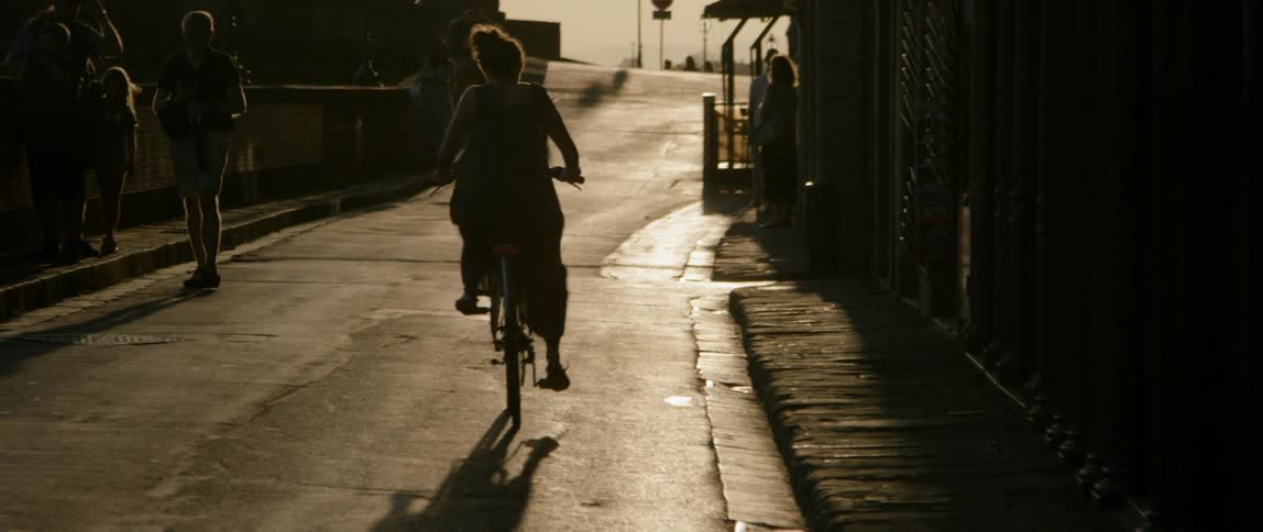 A woman in a dress rides a bike down a building lined street