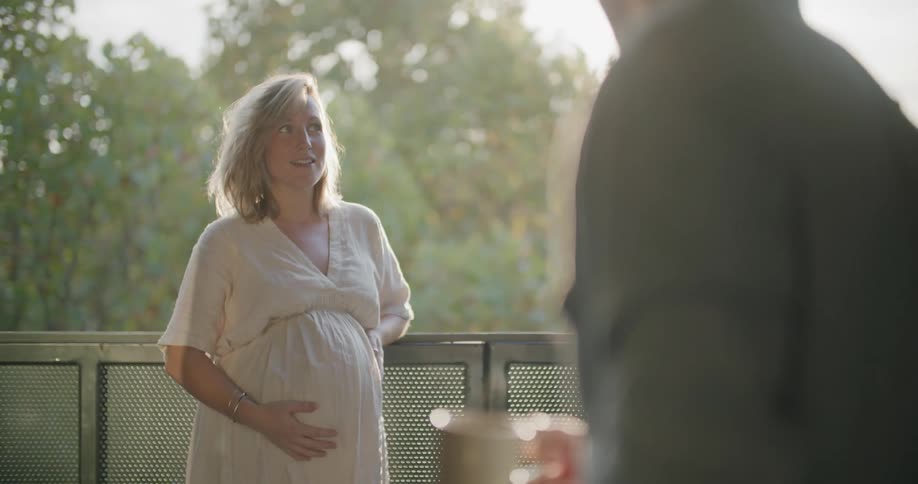Woman strokes her pregnant abdomen while she and spouse have coffee on patio
