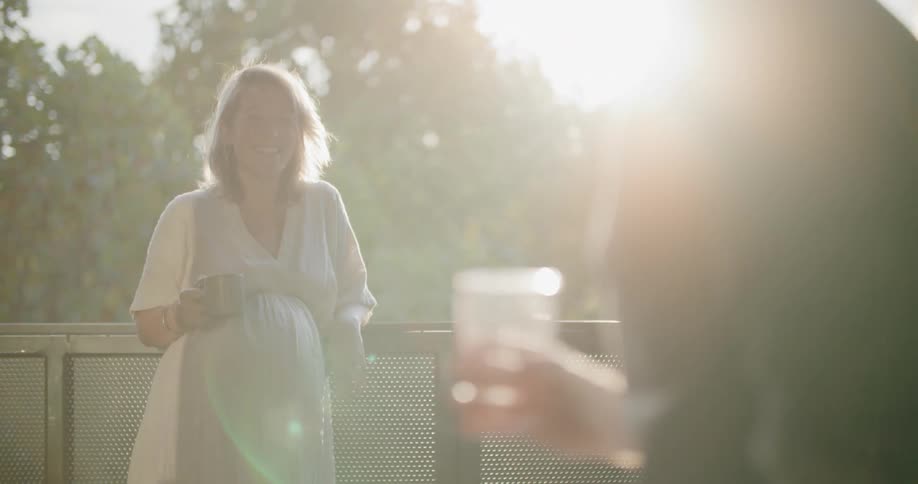 A woman touches her pregnant stomach while laughing and talking to a man on a patio outside