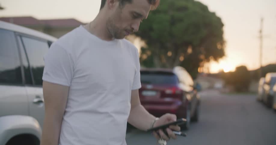 Man with white t shirt and jeans uses smartphone next to cars in the street at sunset