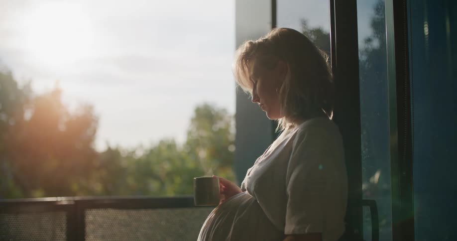 Blonde pregnant woman with a white dress drinks a cup of tea and holds her belly