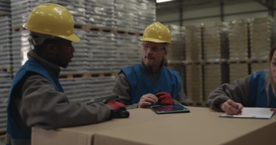 A female worker in a warehouse writes on a clipboard as an older male colleage uses a tablet and a third coworker speaks to them while holding a tool