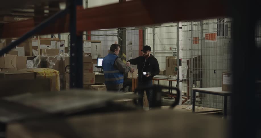 A man with a vest taking a clipboard and a box to another man with a scanner in a warehouse