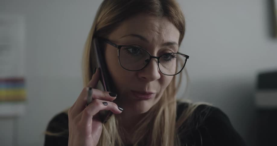 Blonde woman with glasses and a black sweater talks on the smartphone in the office