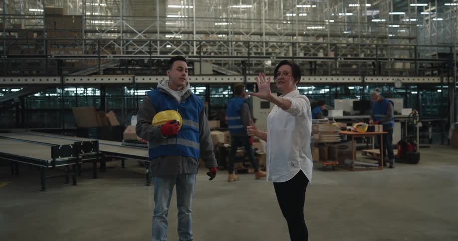 A woman in a white shirt showing a man with a vest and gloves around in a warehouse with lights