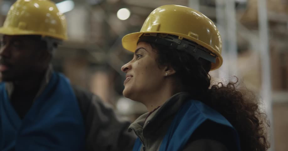 A man and a woman with hardhats and vests in a warehouse with shelves in the background