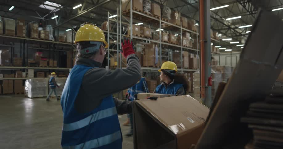 A warehouse worker looks through a stack of collapsed boxes while greeting his coworkers as the pass by